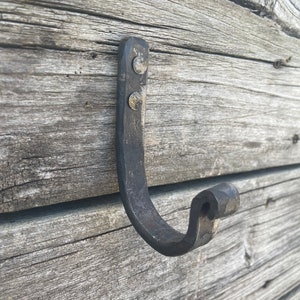 May include: Hand-forged iron hook with a curved design, mounted on weathered wooden planks. The hook is dark gray and secured with two silver rivets, showcasing a rustic aesthetic. Ideal for hanging items.
