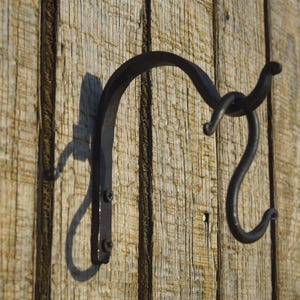 May include: A black, hand-forged iron hook mounted on a weathered wooden surface. The hook has a curved design with a looped detail and a downward-facing hook. The wood has a natural, aged appearance.
