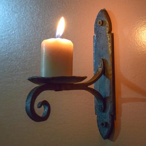 May include: A lit candle in a rustic, wrought-iron wall sconce. The candle is ivory-colored, and the sconce has a decorative, scrolled arm. The sconce is mounted on a dark, textured metal plate, creating a warm, inviting ambiance.