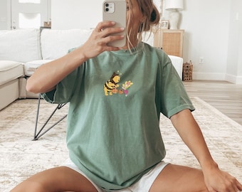 Op de afbeelding: Een vrouw draagt een groen T-shirt met een cartoon-bij-afbeelding, een witte broek en witte sokken met zwarte strepen. Ze zit op een houten vloer en houdt een telefoon voor haar gezicht.