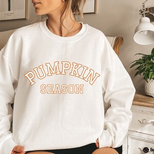 May include: White sweatshirt with the words "PUMPKIN SEASON" in orange block letters. The sweatshirt is a crew neck style. The text is in a collegiate font. The overall theme is autumn and fall.