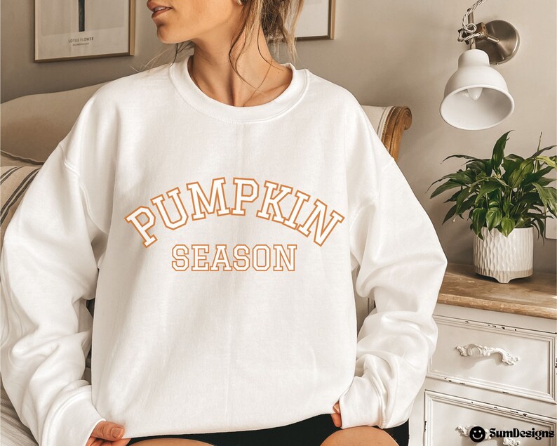 May include: White sweatshirt with the words "PUMPKIN SEASON" in orange block letters. The sweatshirt is a crew neck style. The text is in a collegiate font. The overall theme is autumn and fall.