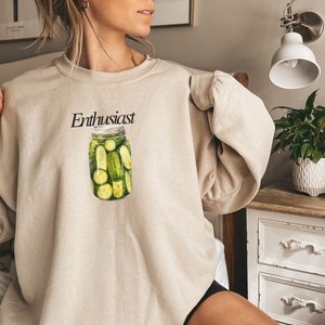 Op de afbeelding: Een beige sweatshirt met een afbeelding van een pot met augurken en het woord "Enthusiast" erop gedrukt.
