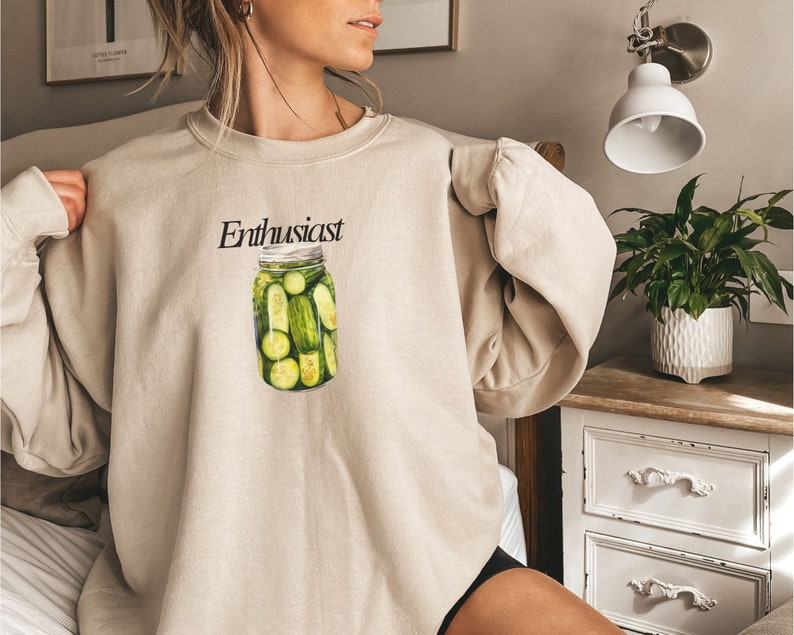 Op de afbeelding: Een beige sweatshirt met een afbeelding van een pot met augurken en het woord "Enthusiast" erop gedrukt.