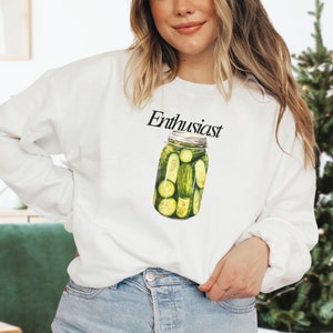 Op de afbeelding: Witte trui met de tekst "Pickle Enthusiast" en een afbeelding van een pot met augurken.