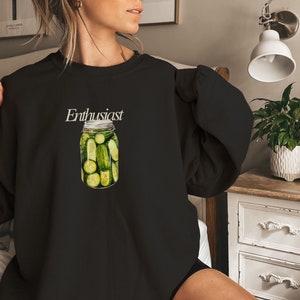 Op de afbeelding: Zwart sweatshirt met de tekst "Enthusiast" en een afbeelding van een glazen pot gevuld met groene augurken.