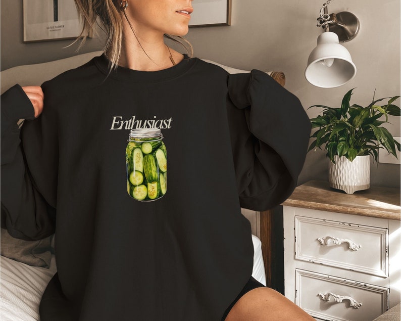 Op de afbeelding: Zwart sweatshirt met de tekst "Enthusiast" en een afbeelding van een glazen pot gevuld met groene augurken.