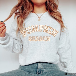 May include: A light gray sweatshirt with the words "PUMPKIN SEASON" in orange block letters. The sweatshirt is a crew neck style and is made of a soft material. The text is in a collegiate style font. The sweatshirt is a fall-themed item.
