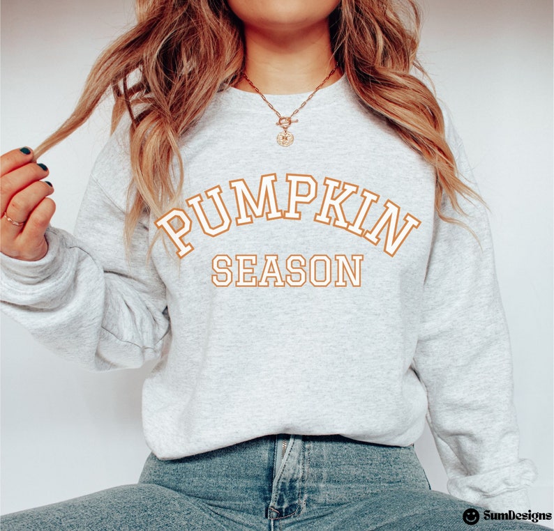 May include: A light gray sweatshirt with the words "PUMPKIN SEASON" in orange block letters. The sweatshirt is a crew neck style and is made of a soft material. The text is in a collegiate style font. The sweatshirt is a fall-themed item.