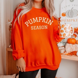 May include: Bright orange sweatshirt with the text "PUMPKIN SEASON" in white. A white mug with orange pumpkin designs is held in the right hand. The background includes pumpkins and fall decor, suggesting a seasonal theme.