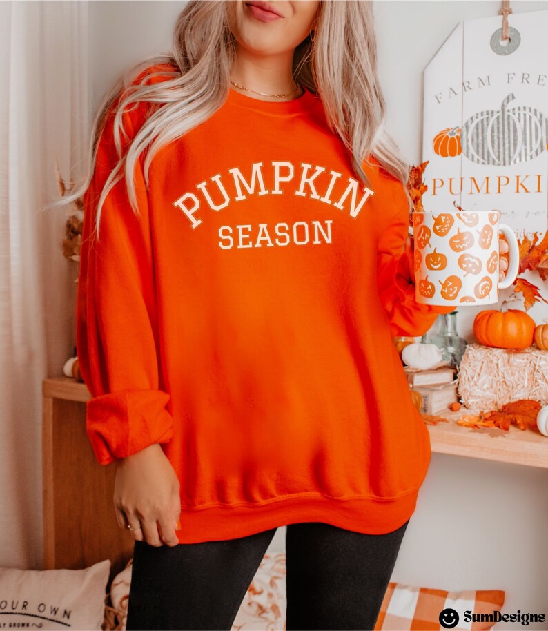 May include: Bright orange sweatshirt with the text "PUMPKIN SEASON" in white. A white mug with orange pumpkin designs is held in the right hand. The background includes pumpkins and fall decor, suggesting a seasonal theme.