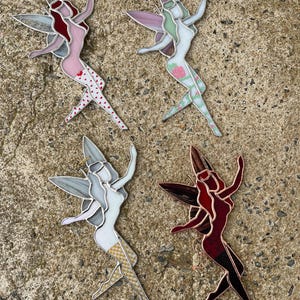 May include: Four stained glass fairy figurines in various colors. One fairy has pink hair and a pink dress with white hearts. Another has green hair and a floral dress. The other two fairies have white and red dresses.