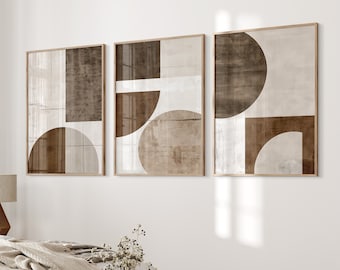 May include: Three framed abstract art prints with geometric shapes in shades of brown and white. The prints are hung on a white wall above a bed with a beige blanket.