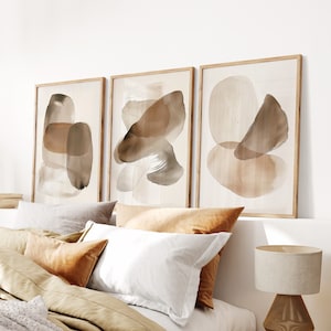 May include: Three framed abstract art prints with brown and beige watercolor shapes on a white background. The prints are hanging on a white wall above a bed with brown and beige bedding.