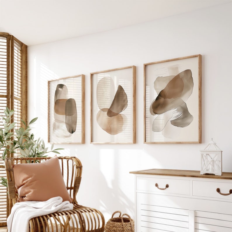 Puede incluir: Tres impresiones de arte abstracto enmarcadas con formas de acuarela marrones y beige sobre un fondo blanco. Las impresiones est&aacute;n colgadas en una pared blanca sobre una c&oacute;moda blanca con tiradores de cajones marrones.