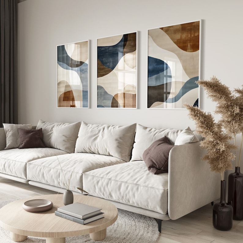 Pode incluir: Tr&ecirc;s impress&otilde;es de arte abstrata com fundo branco e formas azuis e marrons. As impress&otilde;es s&atilde;o emolduradas em branco e penduradas em uma parede branca acima de um sof&aacute; branco.