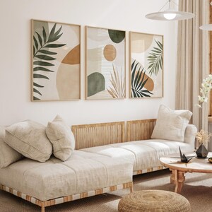 May include: Three abstract art prints with a neutral color palette of beige, brown, and green. The prints feature stylized leaves and geometric shapes. The prints are hanging on a white wall above a light beige couch with wooden slats.