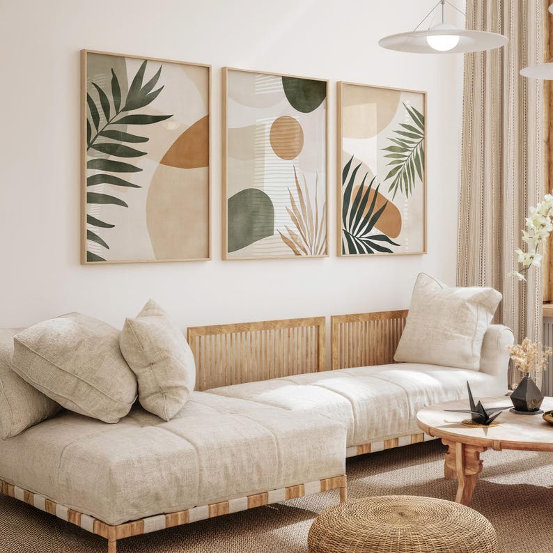 May include: Three abstract art prints with a neutral color palette of beige, brown, and green. The prints feature stylized leaves and geometric shapes. The prints are hanging on a white wall above a light beige couch with wooden slats.