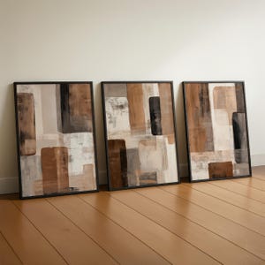 May include: Three framed abstract art prints. Each print features a collage of rectangular shapes in shades of brown, beige, grey, and black. The frames are black, and the artwork leans against a white wall on a wooden floor.