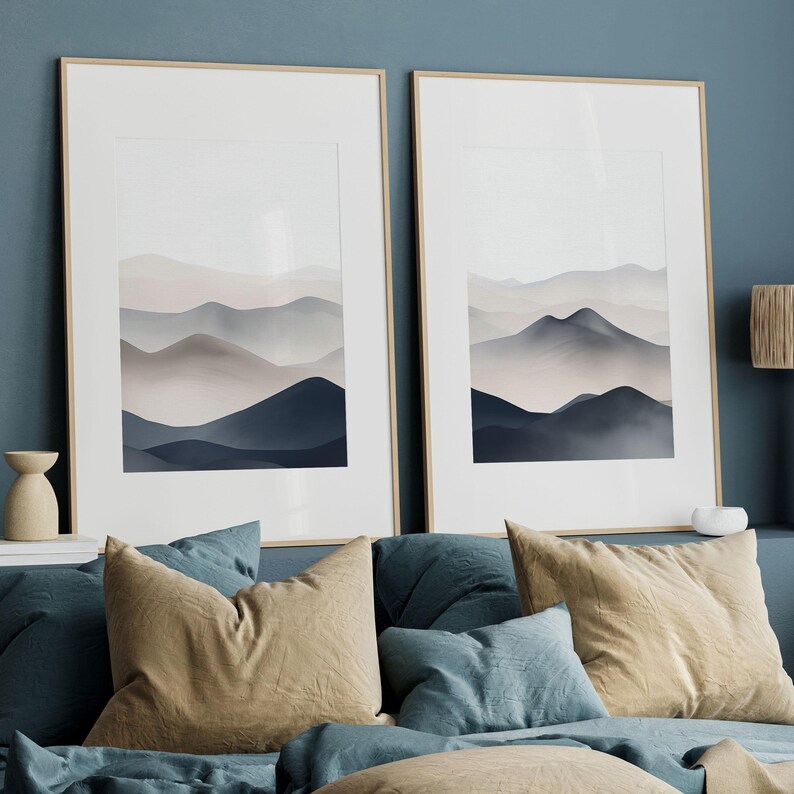 Puede incluir: Dos impresiones enmarcadas de paisajes abstractos con una paleta de colores apagados de azul, gris y blanco. Las impresiones representan una cordillera estilizada con un efecto atmosf&eacute;rico brumoso. Las impresiones est&aacute;n colgadas en una pared azul sobre una cama con almohadas marrones y azules.