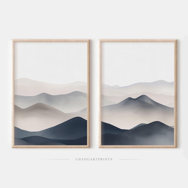 Puede incluir: Dos impresiones enmarcadas de paisajes abstractos de acuarela con una paleta de colores apagados de azul, gris y beige. Las impresiones presentan una serie de colinas y monta&ntilde;as superpuestas.