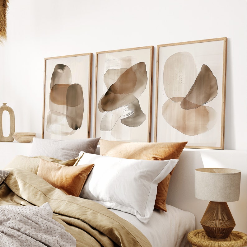 Puede incluir: Tres impresiones de arte abstracto enmarcadas con formas de acuarela marrones y beige sobre un fondo blanco. Las impresiones est&aacute;n colgadas en una pared blanca sobre una cama con ropa de cama marr&oacute;n y beige.