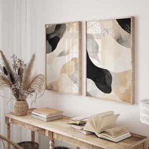 Op de afbeelding: Twee ingelijste abstracte kunstprints met zwart en beige aquarel-achtige ontwerpen. De prints hangen aan een witte muur boven een houten tafel met boeken en een vaas met gedroogde bloemen.