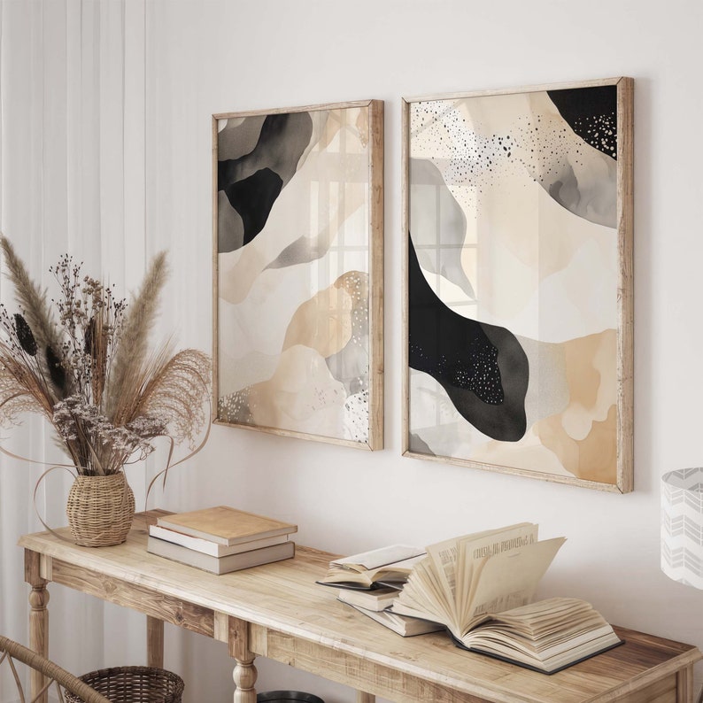 Op de afbeelding: Twee ingelijste abstracte kunstprints met zwart en beige aquarel-achtige ontwerpen. De prints hangen aan een witte muur boven een houten tafel met boeken en een vaas met gedroogde bloemen.
