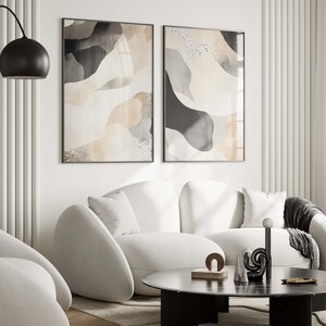 Op de afbeelding: Twee abstracte kunstprints met zwarte en witte vormen op een beige achtergrond. De prints zijn zwart ingelijst en hangen aan een witte muur boven een witte bank.
