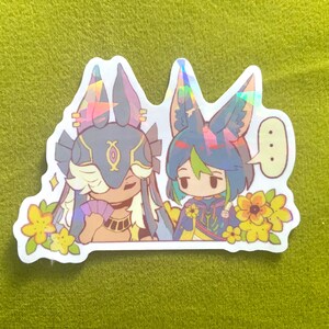 May include: A holographic sticker featuring two anime-style characters with animal ears and floral accents. One character holds a fan, and the other has a speech bubble. The sticker has a white border and is set against a green background.