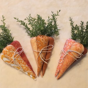 May include: Four handmade fabric carrots with green leafy tops. The carrots are made of orange and yellow patterned fabric and are tied with twine.