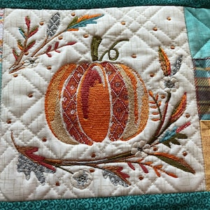 May include: A quilted fabric square with an embroidered pumpkin at the center. The pumpkin is orange and has detailed stitching. Surrounding the pumpkin are embroidered leaves and flowers in fall colors. The background is a cream color with a diamond pattern.