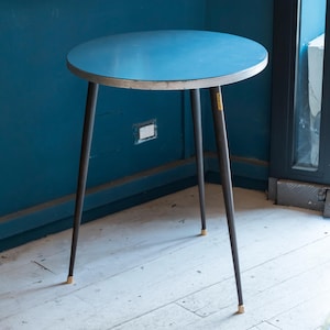 Può includere: Un tavolino rotondo con piano blu e tre gambe marrone scuro. Il tavolo ha un bordo chiaro e piccoli piedini dorati. Il tavolo è alto circa 61 cm.