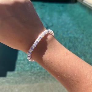 May include: A white and purple beaded bracelet with a simple design. The bracelet is made of small, round beads that are strung together on a clear elastic cord.