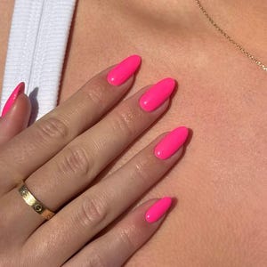 Puede incluir: Un anillo de oro con un motivo repetitivo de cabezas de tornillo, usado en un dedo con uñas largas de forma ovalada pintadas con un esmalte rosa neón brillante y vibrante. El anillo presenta un acabado metálico pulido y un diseño plano.