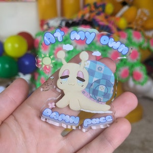 May include: A clear acrylic charm featuring a cartoon snail with a blue and white checkered shell. The snail is smiling and has pink cheeks. The charm has the text "At my own damn pace" around the edge.