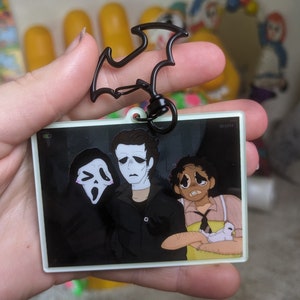 May include: A white acrylic keychain featuring a black and white illustration of three characters from a horror movie. The characters are Ghostface, Michael Myers, and a young woman holding a white bunny. The keychain has a black bat-shaped clip.