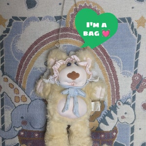 Op de afbeelding: Een pluche teddybeer met een roze strik en een blauw lint, ontworpen als een handtas met een zilveren kettingband. De beer hangt aan een wandkleed met een groene hartvormige spraakballon die zegt "Ik ben een tas".