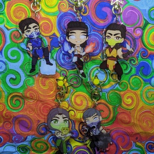 Peut inclure: Cinq porte-clés de personnages de dessins animés colorés avec des breloques en forme d'étoile dorée. Les personnages sont tous masculins et ont des cheveux et des vêtements de couleurs différentes. Les porte-clés sont sur un fond coloré.
