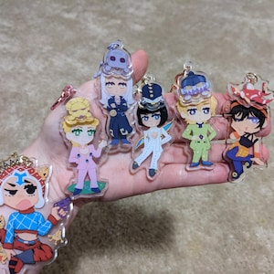Puede incluir: Una colección de seis llaveros de acrílico transparente con personajes de anime estilizados. Un llavero muestra a un personaje con una camisa de patrón azul y el texto MISTAAAA. Otros personajes visten atuendos en rosa, azul oscuro, blanco, verde y negro.