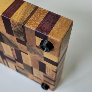 May include: A wooden chopping board with a mosaic pattern of different coloured woods. The board has black rubber feet on the bottom.