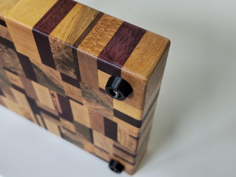 May include: A wooden chopping board with a mosaic pattern of different coloured woods. The board has black rubber feet on the bottom.
