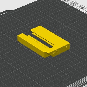 May include: A yellow plastic 3D printed object with the text "NM.92" on the side. The object is designed to hold a small item, such as a key or a piece of jewelry.