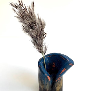 May include: A small, blue ceramic vase with a unique, asymmetrical design. The vase has a textured, speckled finish with hints of gold and orange. A single dried grass stalk is placed inside the vase.
