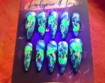 May include: Set of ten press-on nails with a jellyfish design. The nails are a light blue color with a white marbled effect. The jellyfish are painted in a bright green and purple color. The nails are arranged in two rows of five. The text "Larkspur & Ivy" and "Press-on Nails" is printed on the card. The text "@larkspur.and.ivy" is printed on the bottom of the card.