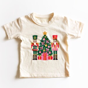 May include: A cream-colored short-sleeve t-shirt featuring a central graphic of a green Christmas tree decorated with pink ornaments and bows, topped with a gold star, flanked by two nutcracker figures and red gift boxes.