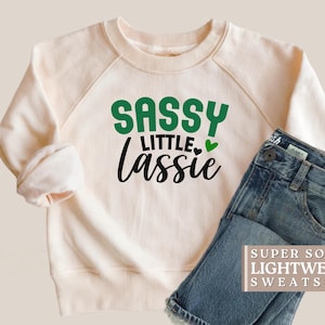 May include: Cream-colored sweatshirt with the text "SASSY LITTLE Lassie" in green and black, with a green heart. Paired with blue denim jeans. The sweatshirt has the words "SUPER SOFT LIGHTWEIGHT SWEATSHIRT".