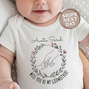 May include: White baby onesie with the text "Auntie Sarah, Will You Be My Godmother?" in script font. The design includes a floral wreath with a hand-holding graphic. A circular sticker reads "Ships Next Day".