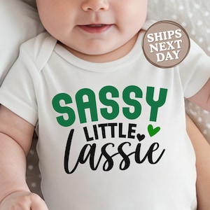 May include: White baby bodysuit with the text "SASSY LITTLE LASSIE" in green and black lettering, with a small green heart. The bodysuit has short sleeves and a round neckline. A circular sticker reads "SHIPS NEXT DAY".