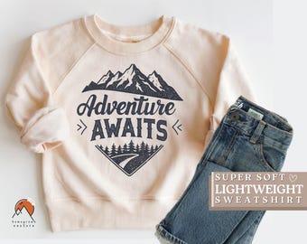 May include: A light beige sweatshirt with a graphic design of mountains and trees. The text "Adventure Awaits" is printed on the sweatshirt. The sweatshirt is super soft and lightweight.
